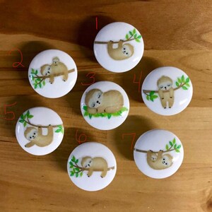 May include: Seven white ceramic knobs with sloth designs. Each knob features a sloth on a branch with green leaves. The sloths are in various poses, including sleeping, hanging, and holding a baby sloth.