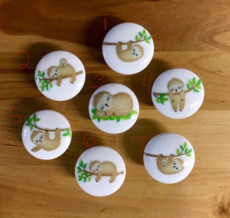 May include: Seven white ceramic knobs with sloth designs. Each knob features a sloth on a branch with green leaves. The sloths are in various poses, including sleeping, hanging, and holding a baby sloth.
