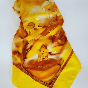 Op de afbeelding: Een gele zijden sjaal met een bruin en oranje ontwerp van zonnebloemen en een klein dier. De sjaal heeft een gele rand en het woord "Feiber" staat in het midden.