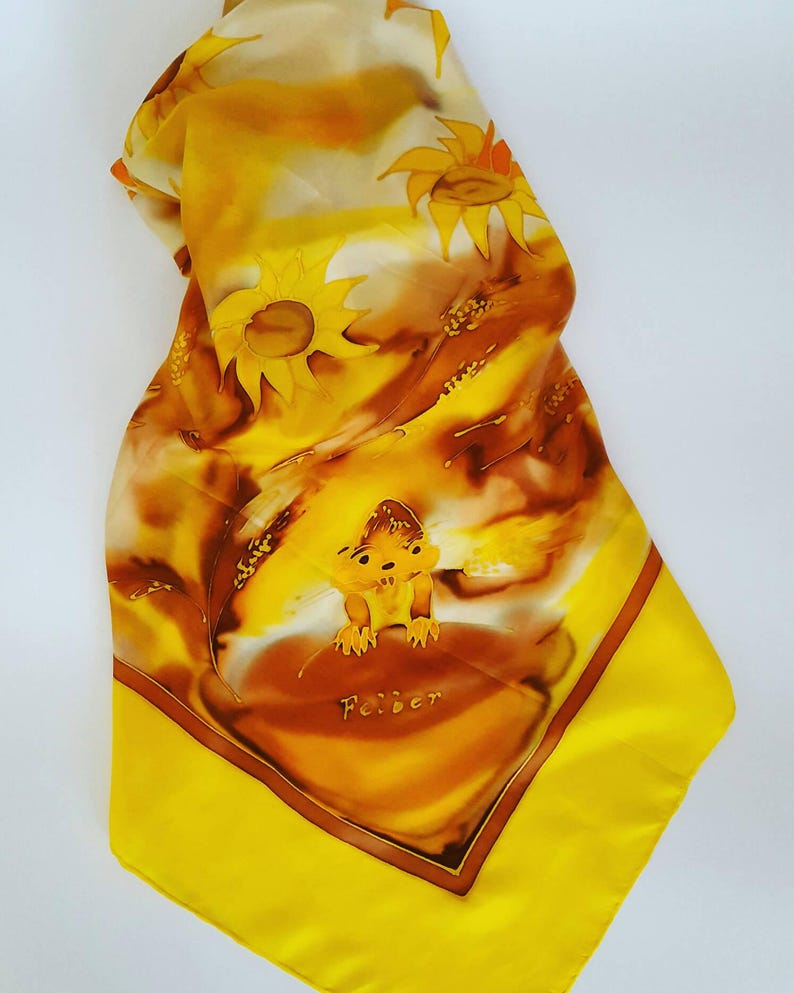 Op de afbeelding: Een gele zijden sjaal met een bruin en oranje ontwerp van zonnebloemen en een klein dier. De sjaal heeft een gele rand en het woord "Feiber" staat in het midden.