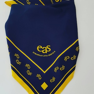 Può includere: Un bandana blu navy con bordo giallo e il testo "eas EUROPEAN AVIA SERVICE" al centro. Il bordo è decorato con il testo "eas" ripetuto più volte.