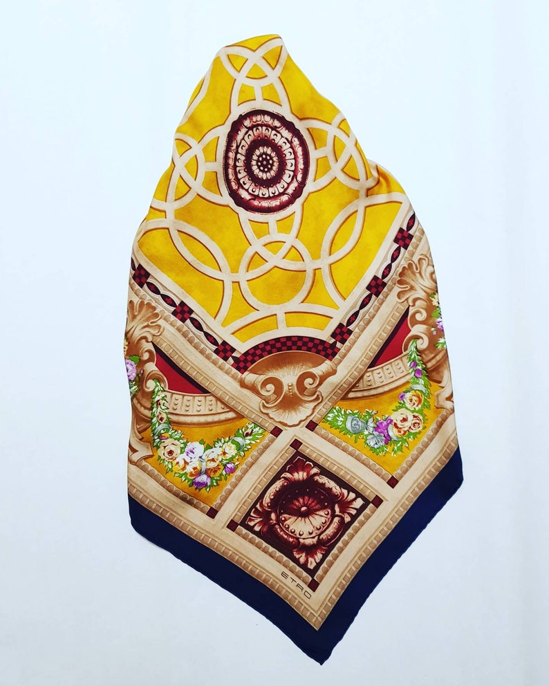 May include: A yellow, red and blue silk scarf with a geometric pattern and floral accents. The scarf features a central design of interlocking circles and a smaller floral design in the bottom corner. The brand name "ETRO" is printed in the bottom corner.