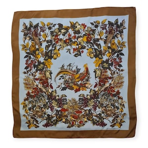 Puede incluir: Pañuelo cuadrado de seda con un centro azul claro con un diseño de faisán y pájaros rodeado de follaje en tonos amarillos, marrones y rojos. El borde es marrón liso.