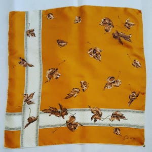 May include: A square silk scarf with a brown and white leaf pattern on a mustard yellow background. The scarf has a white border with a brown and white geometric pattern. The signature "Clesco" is in the bottom right corner.