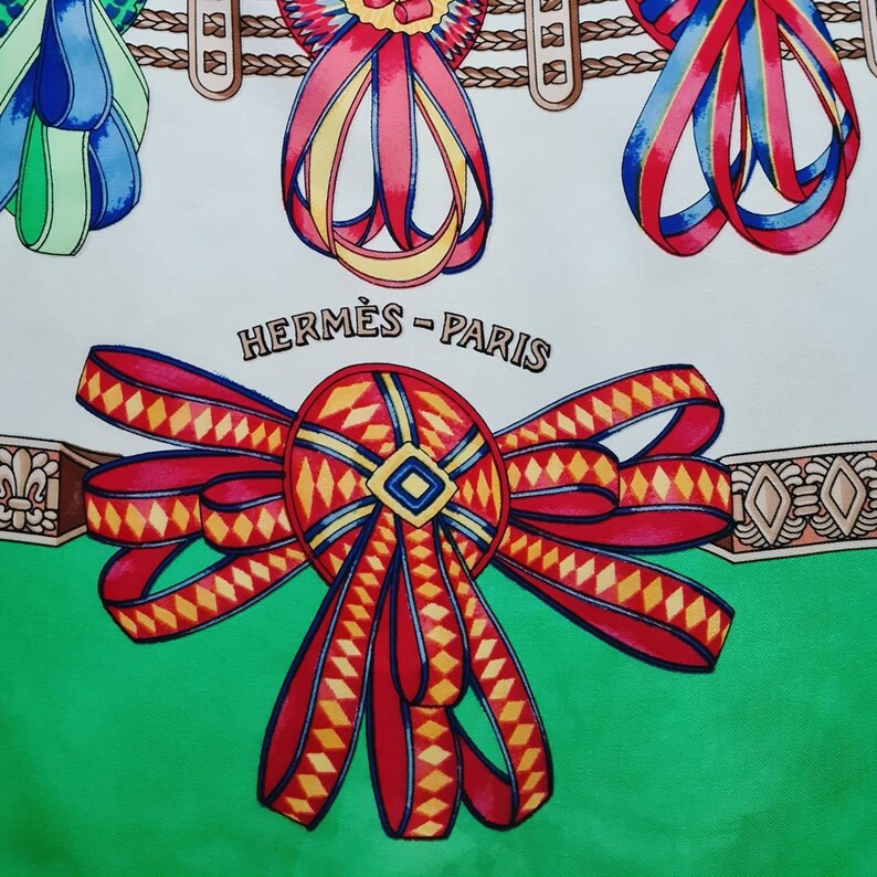 May include: A red, yellow, and white bow with a diamond pattern on a green background. The text "HERM&Egrave;S - PARIS" is printed in black above the bow.
