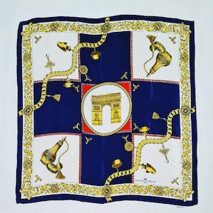 May include: A blue and white silk scarf with a gold chain design and a depiction of the Arc de Triomphe in Paris. The scarf also features a gold and red design with the text "Parfums Creatifs" in the bottom right corner.