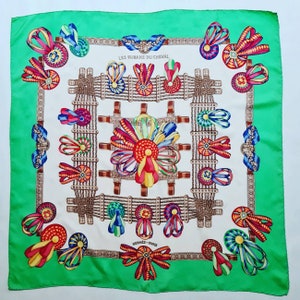 May include: A white silk scarf with a green border. The scarf features a repeating pattern of colorful ribbons and rosettes. The text "LES RUBANS DU CHEVAL" and "HERMES - PARIS" are printed on the scarf.