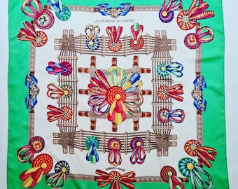 May include: A white silk scarf with a green border. The scarf features a repeating pattern of colourful ribbons and rosettes. The text "LES RUBANS DU CHEVAL" and "HERMES - PARIS" are printed on the scarf.