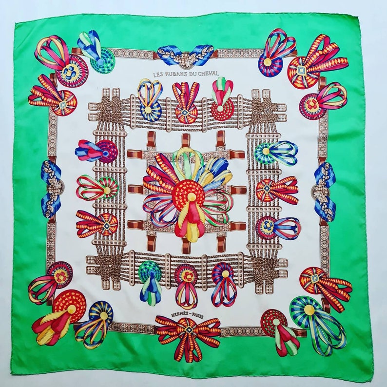 May include: A white silk scarf with a green border. The scarf features a repeating pattern of colorful ribbons and rosettes. The text "LES RUBANS DU CHEVAL" and "HERMES - PARIS" are printed on the scarf.