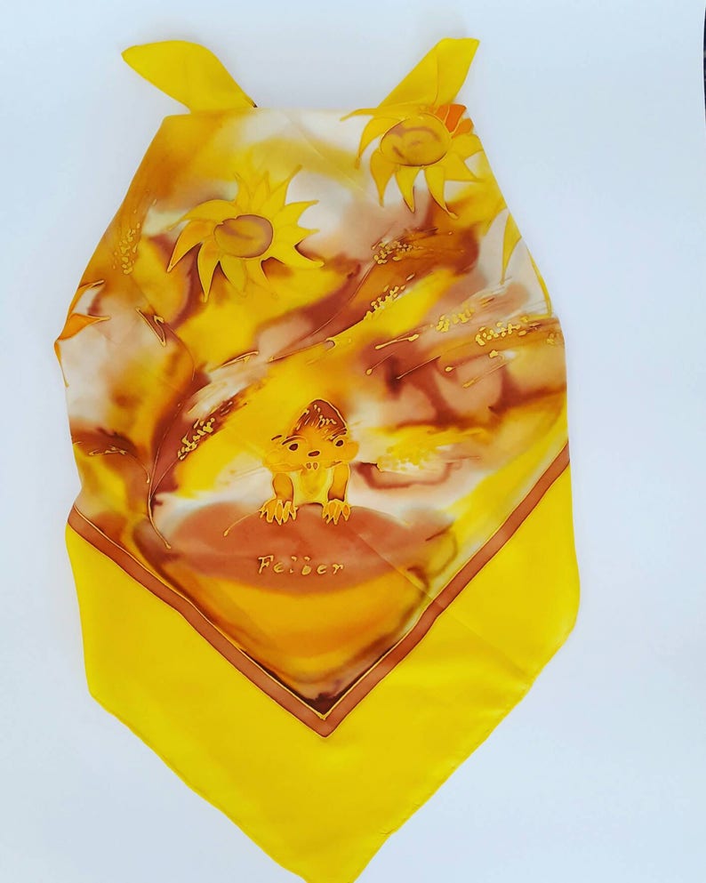 Op de afbeelding: Gele zijden sjaal met een handgeschilderd ontwerp van zonnebloemen, tarwe en een klein dier. De sjaal heeft een bruine rand en de tekst "Feiber" is in het midden gedrukt.
