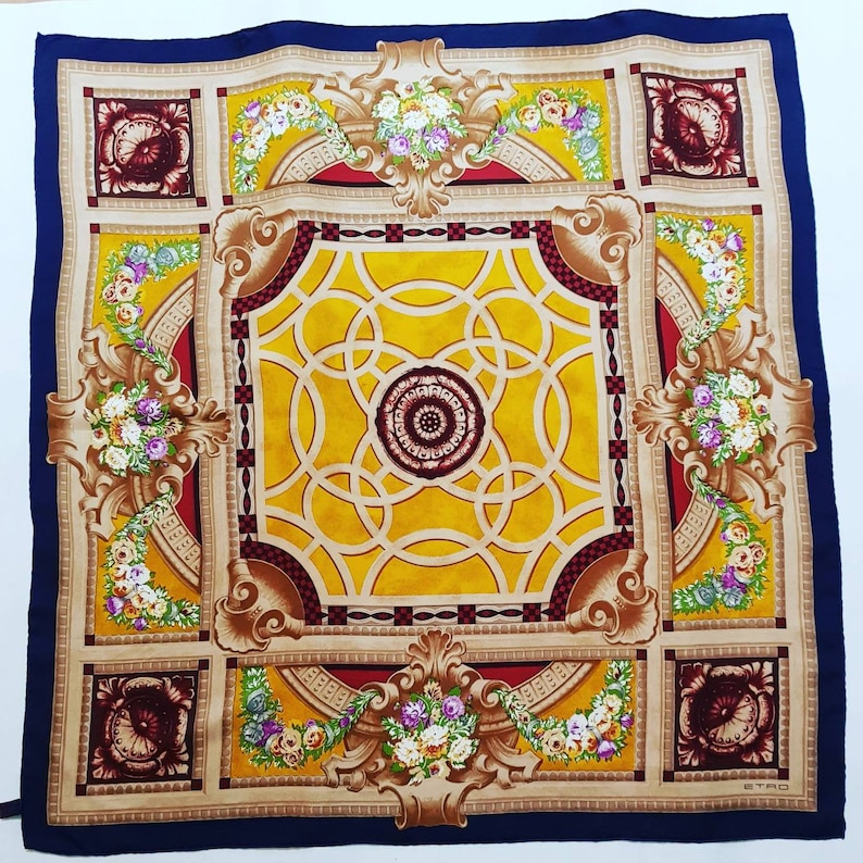 May include: A blue silk scarf with a gold and red patterned design. The pattern features a central ornate design with a circular motif and floral accents. The text "ETRO" is visible in the bottom right corner.