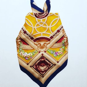 May include: A yellow, red and blue silk scarf with a geometric pattern and floral accents. The scarf features a gold and brown design with a central motif of a stylised flower. The scarf is tied in a knot at the top and has the word "ETRO" printed in the bottom corner.