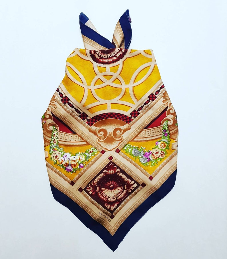 May include: A yellow, red and blue silk scarf with a geometric pattern and floral accents. The scarf features a gold and brown design with a central motif of a stylised flower. The scarf is tied in a knot at the top and has the word "ETRO" printed in the bottom corner.