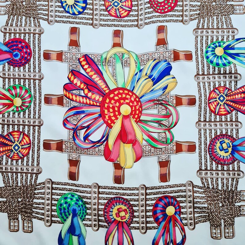 May include: A white fabric with a repeating pattern of brown rope, colorful rosettes, and ribbons. The rosettes are in shades of red, blue, green, and yellow. The ribbons are in shades of red, blue, green, and yellow.