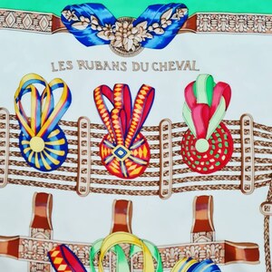 May include: A white fabric with a green border and a repeating pattern of brown rope, brown leather straps, and colorful ribbons tied in bows. The text "LES RUBANS DU CHEVAL" is printed in black at the top of the fabric.