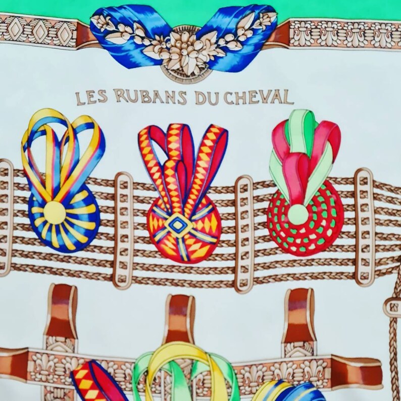 May include: A white fabric with a green border and a repeating pattern of brown rope, brown leather straps, and colorful ribbons tied in bows. The text "LES RUBANS DU CHEVAL" is printed in black at the top of the fabric.