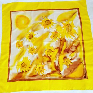 Op de afbeelding: Een gele zijden sjaal met een witte achtergrond met een geschilderd ontwerp van zonnebloemen en een zon. De sjaal heeft een bruine rand en het woord "Feiler" is gedrukt in de rechterbenedenhoek.