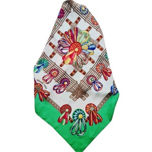 May include: A white silk scarf with a green border. The scarf features a repeating pattern of colorful ribbons and bows tied around medallions. The text "HERMES - PARIS" is printed in the lower left corner.