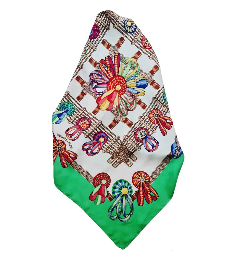 May include: A white silk scarf with a green border. The scarf features a repeating pattern of colorful ribbons and bows tied around medallions. The text "HERMES - PARIS" is printed in the lower left corner.