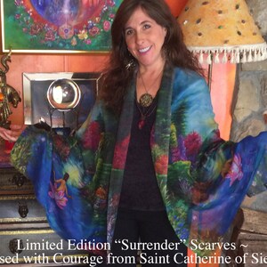 May include: A woman wearing a blue silk scarf with a colorful floral and forest scene print. The scarf is called "Surrender" and is a limited edition. The text "Blessed with Courage from Saint Catherine of Sienna" is printed below the scarf.