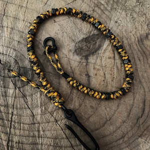 May include: A black and yellow paracord keychain with a black metal hook. The keychain is made with a braided paracord and has black metal beads.