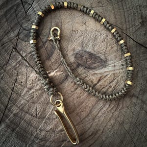Könnte beinhalten: Ein grüner Paracord-Schlüsselanhänger mit Messingbeschlägen. Der Schlüsselanhänger hat einen Messinghaken und eine Reihe von Messingperlen.