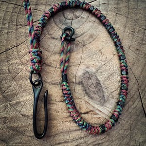 May include: A multi-colored paracord lanyard with a black metal clasp and hardware. The lanyard features a rainbow pattern of red, yellow, green, and blue threads, woven together. The hardware includes a hook and a series of square-shaped beads.