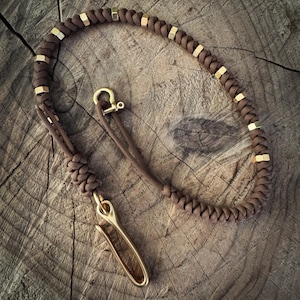 May include: A brown paracord leash with gold-colored metal accents and a matching clasp. The leash is knotted and braided, with evenly spaced gold-colored metal pieces. The clasp is a gold-colored metal hook.