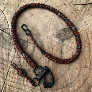 May include: A brown paracord lanyard with black metal hardware. The lanyard features a series of dark gray metal hex nuts and a black carabiner-style clasp. The cord is tightly woven and appears durable, suitable for outdoor use.