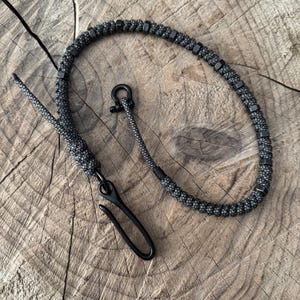 May include: A black and gray paracord lanyard with a black metal clasp and hardware. The lanyard features a woven design with black metal beads. The hook is curved and the lanyard is lying on a wooden surface.
