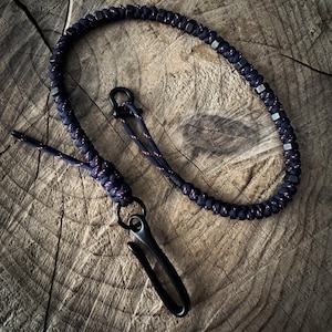 May include: A dark blue lanyard with a black metal clasp and hardware. The lanyard is made of braided cord with small, square metal accents. The background is a textured, wooden surface.