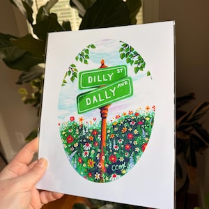 May include: A rectangular art print featuring an oval illustration of two green street signs labeled DILLY ST and DALLY AVE. The signs are mounted on a brown post amidst a vibrant field of colorful wildflowers under a light blue sky.