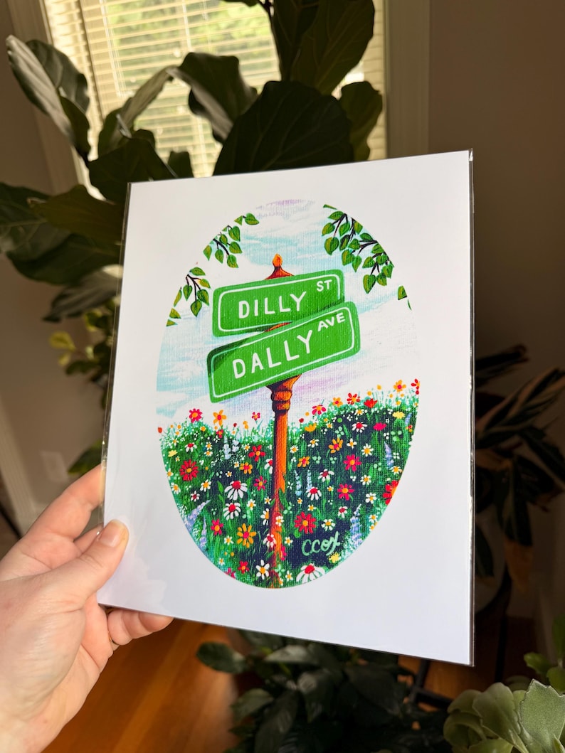 May include: A rectangular art print featuring an oval illustration of two green street signs labeled DILLY ST and DALLY AVE. The signs are mounted on a brown post amidst a vibrant field of colorful wildflowers under a light blue sky.