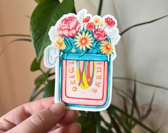 Pode incluir: Um autocolante colorido com um arranjo floral num frasco. O frasco é azul e rosa com as palavras "ORIGI SARDI". As flores incluem peónias vermelhas, margaridas amarelas e flores laranja. O autocolante é segurado por uma mão.