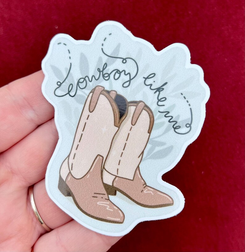 May include: A sticker featuring a pair of brown cowboy boots and the text "So cowboy like me" in a cursive font on a light blue background with a leafy pattern.