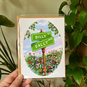 May include: A rectangular greeting card in a clear plastic sleeve featuring an oval illustration of two green street signs labeled DILLY ST and DALLY AVE on a brown post, set against a vibrant meadow of colorful wildflowers and a blue sky.