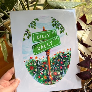 May include: A rectangular art print featuring an oval illustration of two green street signs labeled Dilly St and Dally Ave. The signs are mounted on a brown post surrounded by a vibrant field of colorful wildflowers under a light blue sky.