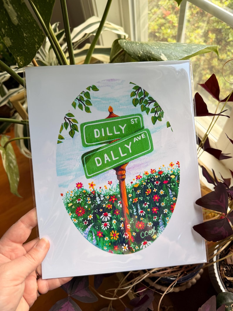 May include: A rectangular art print featuring an oval illustration of two green street signs labeled Dilly St and Dally Ave. The signs are mounted on a brown post surrounded by a vibrant field of colorful wildflowers under a light blue sky.