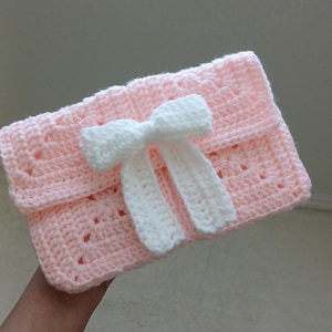 May include: A handmade, light pink crochet clutch with a white bow. The clutch has a folded-over flap and a textured stitch pattern. The white bow is centered on the front, adding a decorative touch. The overall design is soft and feminine.