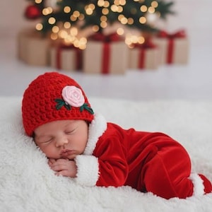 May include: A sleeping baby dressed in a red Santa suit and hat with a pink flower, lying on a white fluffy surface. A Christmas tree with lights and wrapped gifts are in the background.