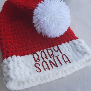 May include: A red crocheted Santa hat with a white fluffy brim and a large white pom-pom. The words "BABY SANTA" are embroidered in red on the brim. Festive holiday apparel.