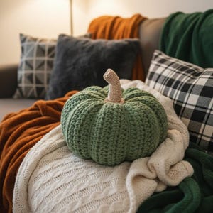 May include: A green crocheted pumpkin with a tan stem sits atop a cream-colored cable-knit blanket. The pumpkin is surrounded by orange, green, and black and white plaid throw blankets and pillows, creating a cozy autumn theme.