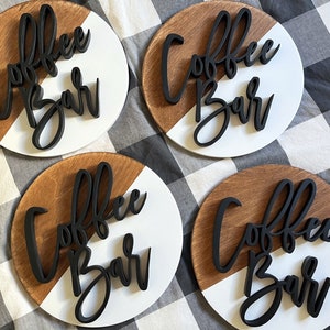 Peut inclure: Quatre pancartes rondes en bois avec un design peint en blanc et brun. Chaque pancarte porte l'inscription "Coffee Bar" en lettres 3D noires.