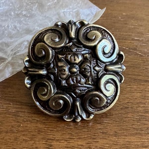 May include: A round, ornate brass knob with a detailed, intricate design. The knob has a central square with a floral pattern and is surrounded by swirling, scroll-like details.