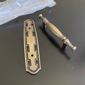 May include: A set of two antique brass cabinet hardware pieces. One is a rectangular plate with an ornate design and the other is a pull handle with a simple design.