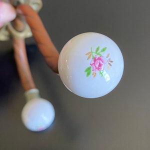 May include: Two white ceramic hooks with a floral design. The hooks are attached to a brown metal base.