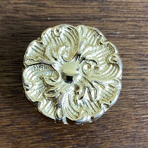 May include: A gold-toned metal knob with a decorative floral design. The knob has a raised, textured surface and a circular shape.