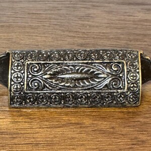 May include: Antique bronze-toned drawer pull with ornate detailing. The rectangular center features a central leaf design, surrounded by intricate scrollwork and circular patterns. The pull is set against a wooden surface.