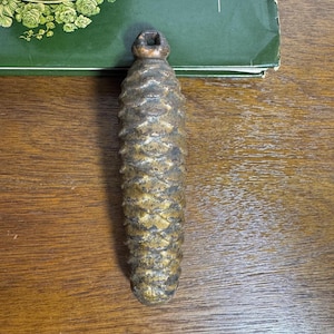 May include: A heavy, cast metal weight shaped like a pinecone with a dark, weathered bronze-toned finish. The object features a textured, overlapping scale pattern and a small, integrated loop at the top for hanging or attachment purposes.