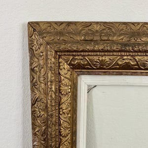 May include: Ornate gold-colored picture frame with detailed floral and scrollwork carvings. The frame has a white interior and is set against a plain white wall. The frame's design suggests a vintage or antique style, suitable for artwork or photographs.